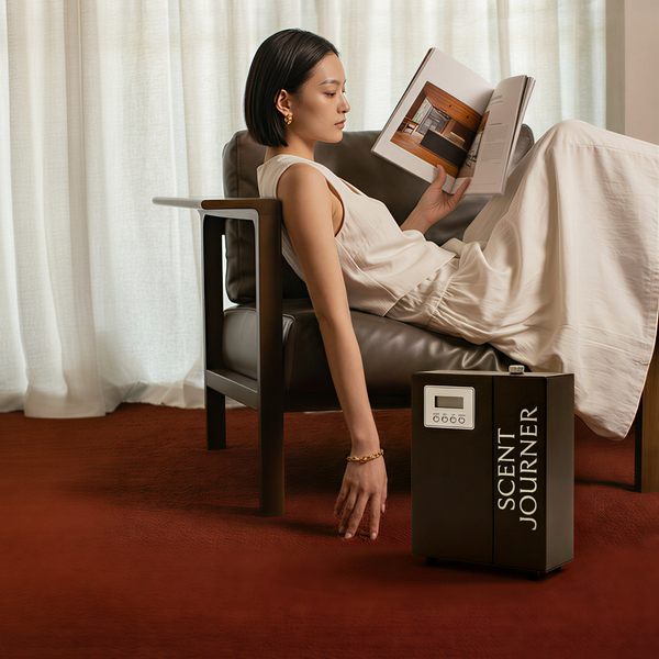 Woman reading a book in a chair next to a 'Scent Journer' diffuser.